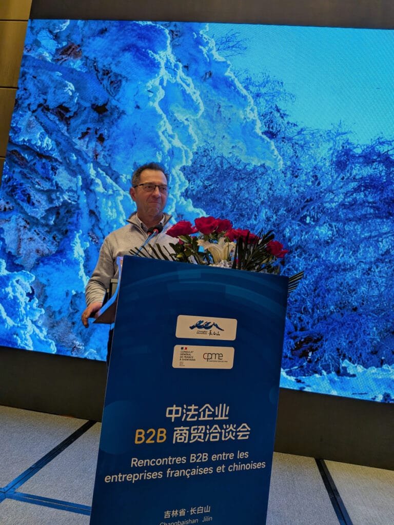 Photo de Christian Gaut en train de faire une conférence en Chine. Il est derrière un pupitre en train de parler et derrière lui on voit un écran immense qui montre un paysage enneigé.